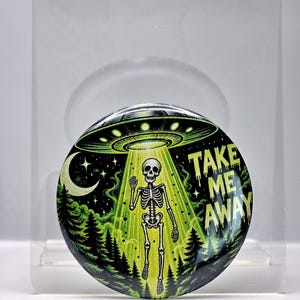 May include: A circular pinback button featuring a graphic illustration of a skeleton being abducted by a green glowing UFO above a forest at night. The design includes a crescent moon and the text TAKE ME AWAY in a stylized, dripping font.