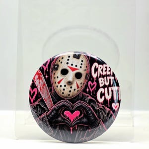 May include: A circular pinback button featuring a hockey mask-wearing figure holding a bloody machete. The figure forms a heart shape with gloved hands. The design includes pink hearts and the text CREEPY BUT CUTE in a dripping, stylized font.