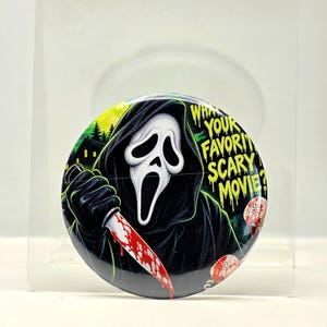 May include: A circular pinback button featuring a graphic illustration of a masked figure in a black hooded robe holding a blood-stained knife. The text "WHATS YOUR FAVORITE SCARY MOVIE?" is printed in yellow, with a green forest background.