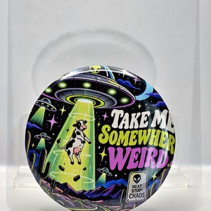 May include: A circular pinback button featuring a colorful illustration of a cow being abducted by a UFO. The text reads "TAKE ME SOMEWHERE WEIRD" in yellow and pink, with a road sign that says "NEXT STOP: CHAOS" and an alien icon.