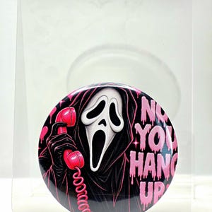May include: A circular pinback button featuring a black-hooded figure with a white ghost mask holding a bright pink telephone receiver. The text 'NO YOU HANG UP' is printed in a dripping pink font on the right side against a dark background.