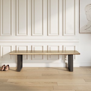 May include: A wooden bench with black metal legs. The bench is made of natural wood with a light brown finish. The bench is sitting on a light wood floor in front of a white wall with wainscoting.