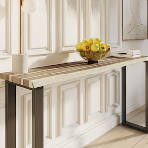 May include: A long, narrow console table with a natural wood top and black metal legs. The table has a live edge design and is decorated with a bowl of yellow apples.