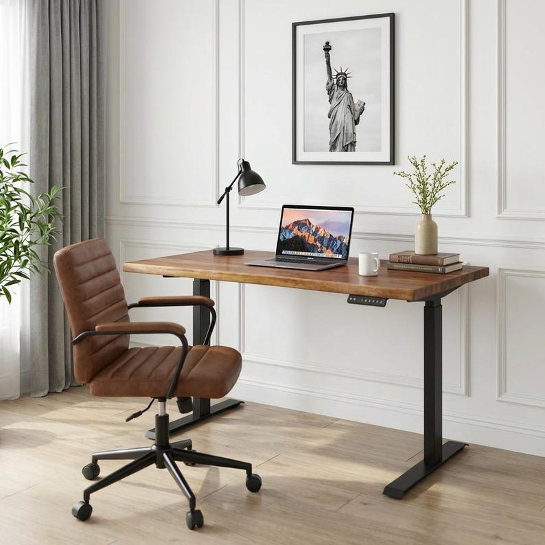May include: A brown leather office chair with a black base and wheels sits in front of a wooden desk with black legs. A laptop, lamp, and framed Statue of Liberty art are on the desk. A vase with greenery and a coffee mug are also on the desk.