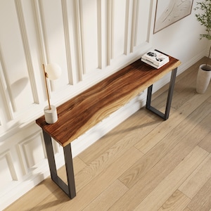 May include: A long, natural wood console table with a live edge and dark metal legs. The table is approximately 72 inches long. A small lamp and books are on the table, which is placed against a white wall.