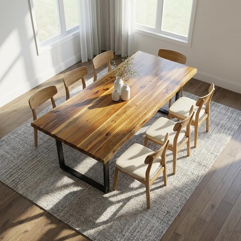 May include: A wooden dining table with a natural finish, supported by a black metal base, surrounded by six wooden chairs with cream-colored upholstered seats. The table is set on a gray patterned rug, with vases and greenery in the center.