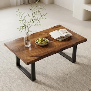 May include: A rectangular wooden coffee table with a natural edge and dark brown finish. It has black metal legs. A clear glass vase with greenery, a bowl of green apples, and an open book are on the table.