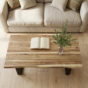 May include: A rectangular wooden coffee table with a natural wood grain finish. The table has black metal legs and a live edge design. An open book and a small vase with greenery are on the table, with a beige sofa in the background.