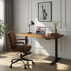 May include: A home office setup featuring a brown wooden desk with black legs, a brown leather office chair, and a laptop. A black and white framed picture of the Statue of Liberty hangs on the wall. A black desk lamp and a vase with greenery are on the desk.