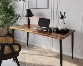 May include: A wooden desk with a live edge top and black metal legs. A laptop, lamp, and a white robot figurine are on the desk. The desk is in a room with a white wall and a plant in the corner.