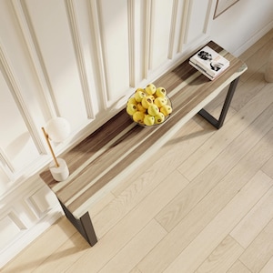 May include: A long, narrow console table with a natural wood top and black metal legs. The table has a live edge design and is decorated with a bowl of yellow fruit.