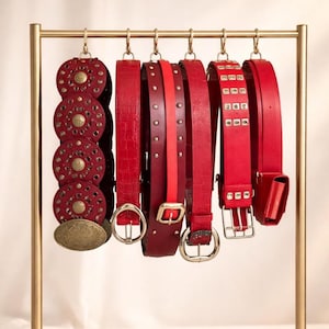 Puede incluir: Una colección de cinturones rojos con varios diseños colgados en un estante dorado. Los cinturones incluyen estilos con tachuelas, grabados y texturas. Algunos tienen hebillas y herrajes dorados, otros elementos decorativos.