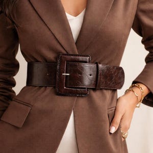 May include: A wide, dark brown belt with a large square buckle cinches the waist of a brown blazer. The belt has a textured appearance. A gold bracelet and ring are visible on the right arm.