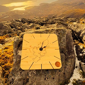 May include: A square wooden clock with a light wood finish, featuring a map outline and black clock hands. The clock is set against a scenic mountain and water backdrop. A small, circular emblem is in the lower right corner.