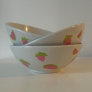 May include: Two white ceramic bowls stacked. Each bowl features a repeating pattern of pink strawberries and watermelon slices with green detailing. The bowls have a smooth, glossy finish. The bowls are likely for serving food.