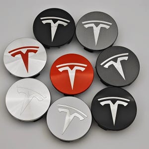 Op de afbeelding: Een verzameling van acht Tesla wielnaafdoppen in verschillende kleuren, waaronder zwart, grijs, rood en zilver. Elke dop heeft het Tesla-logo in een contrasterende kleur, met een geborstelde metalen of effen afwerking.