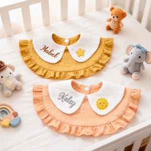 May include: Two baby bibs with white collars and ruffled edges. One is mustard yellow with a star, the other is peach with a sun. Both bibs have a name embroidered on the collar.