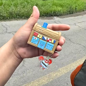 Op de afbeelding: Een bruin rechthoekig etui met een blauwe bovenkant en blauwe accenten, dat op een gebouw lijkt. Er is een sleutelhanger met een stripfiguur bevestigd. De behuizing heeft decoratieve vlaggen en ramen. De sleutelhanger heeft een stripfiguur met rode klauwen.