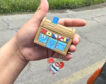 May include: A tan-coloured, rectangular case with a blue top and blue accents, resembling a building. A keychain with a cartoon character is attached. The case has decorative flags and windows. The keychain features a cartoon character with red claws.