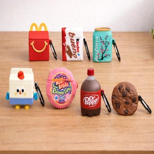 May include: A collection of colorful AirPod cases designed to resemble various food and beverage items. The cases include a red McDonald's Happy Meal, a Kinder Bueno candy bar, an Arizona iced tea can, a milk carton, Hubba Bubba bubble tape, a Dr. Pepper bottle, and a chocolate chip cookie.