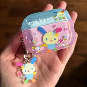 May include: A clear, pink and blue case featuring a cartoon bunny design, with a matching bunny keychain. The text "u-sa-ho-na" is printed on the top. The case is decorated with flowers and a bee.