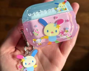 May include: A clear, pink and blue case featuring a cartoon bunny design, with a matching bunny keychain. The text "u-sa-ho-na" is printed on the top. The case is decorated with flowers and a bee.