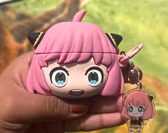 May include: Pink Anya Forger character AirPod case with a matching keychain. The case features a cartoon face with large blue eyes, pink hair, and black accents. The keychain has a small Anya Forger figure.