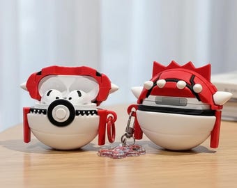 May include: Two white and red AirPod cases are displayed on a wooden surface. One case is open, revealing white earbuds, and the other is closed. The cases have a spherical base and a red top with white accents. A small keychain is attached to the closed case.