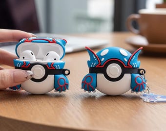 May include: A blue, white, and red Kyogre-themed AirPod case, open to reveal white earbuds. The case is on a wooden surface. A second case is also visible, with a keychain attached.