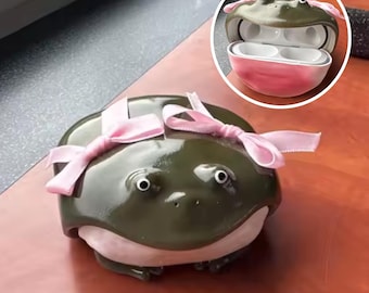 May include: A novelty frog-shaped case for wireless earbuds. The case is dark green with a pink base and two pink bows. The inset image shows the case open, revealing the earbuds inside.