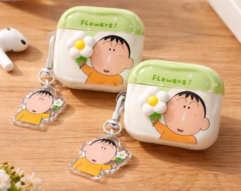 May include: Two white and green AirPod cases featuring a cartoon character holding a flower. The cases are labelled "Flowers!". Each case has a matching keychain with the same character. The background is a light brown wooden surface.