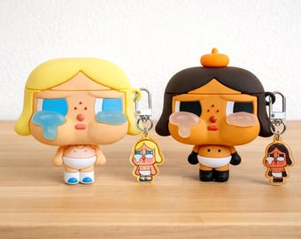 May include: Two cartoon figures with crying faces, one blonde and one brunette, each with a matching keychain. The figures are in a baby-like style, with white nappies and blue tears. The figures are in a baby-like style, with white nappies and blue tears.