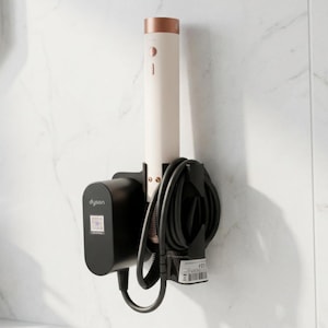 Pode incluir: Um suporte de parede preto para uma ferramenta de modelagem de cabelo, com um dispositivo cilíndrico branco-sujo e topo cor de cobre. O suporte preto inclui uma seção para o adaptador de energia com a marca Dyson e um cabo preto enrolado.
