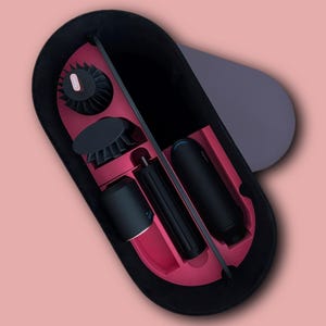 Pode incluir: Um estojo de armazenamento oval com exterior cinza-escuro e interior magenta vibrante. O estojo contém uma ferramenta de modelagem de cabelo preta, um cabo cilíndrico e dois acessórios de escova circulares com cerdas pretas e pequenos detalhes rosa.