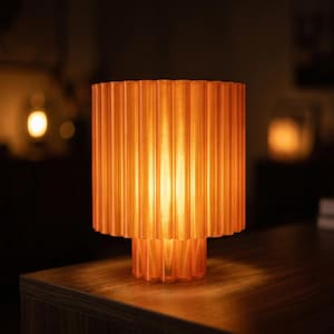 Puede incluir: Una lámpara de mesa de color naranja con una pantalla y una base acanaladas. La lámpara está iluminada, emitiendo un brillo cálido. La lámpara está sobre una superficie de madera.