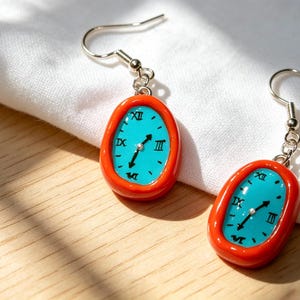 May include: A pair of handmade earrings featuring a clock face design. Each earring has a teal-colored clock face with black Roman numerals and hands, set within a bright orange oval frame. The earrings hang from silver-colored fish hook ear wires.