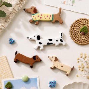 May include: A selection of dachshund dog-shaped magnets in different colours and patterns. One dog wears a yellow sweater with pink polka dots. Other magnets include a black and white spotted dog and a brown dog.
