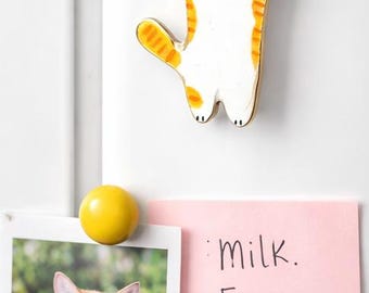May include: A white and orange cat-shaped magnet is displayed on a white refrigerator door. A photo of a cat in a teacup and a pink sticky note with a grocery list are also attached. A yellow round magnet holds the photo and note.