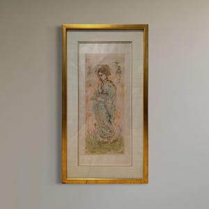 May include: A framed artwork featuring a woman in a kimono. The artwork has a gold frame and depicts a woman in a kimono with a light blue and pink color scheme. The artwork is mounted on a light-colored background.