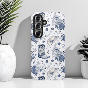 Peut inclure: Une coque de smartphone blanche ornée d'un motif répétitif de bottes de cow-boy bleues, de chapeaux de cow-boy et de compositions florales. Le design comprend diverses fleurs et tiges feuillues dans des tons bleus sur fond blanc.