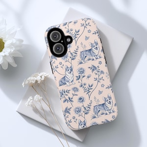 May include: A phone case with a light pink background and a blue and white floral pattern featuring corgis. The case has a black camera cutout.