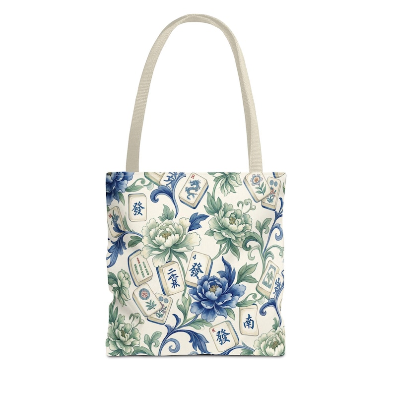 May include: Tote bag with a cream-colored handle and a floral pattern. The pattern features blue and green peonies, leaves, and Mahjong tiles with Chinese characters. The bag is white with a cream-colored handle.