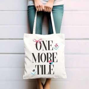 May include: Cream-coloured tote bag with the words "ONE MORE TILE" in large black letters. The bag has a pink bow, a small floral design, and two tile illustrations. The bag is held by a person.