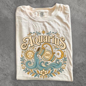 May include: A cream-coloured t-shirt featuring a graphic print of the word Aquarius in a large, stylised font. The design includes a central pitcher pouring water, surrounded by a sun, a crescent moon, floral elements, and stars in blue and gold.