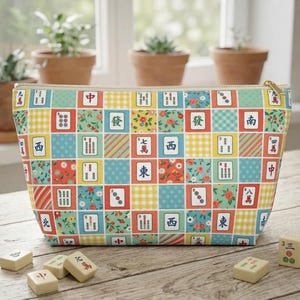 Può includere: Un astuccio colorato con cerniera con un motivo di tessere di Mahjong in vari colori. Il design include elementi floreali e geometrici. Diverse tessere di Mahjong sono sparse davanti all'astuccio su una superficie di legno.