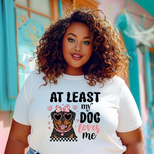 May include: White t-shirt with a black and white illustration of a Rottweiler wearing pink sunglasses and a pink bow. The text "At least my dog loves me" is printed on the shirt.