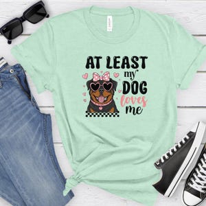 May include: A light green t-shirt with the text "At Least My Dog Loves Me" in pink and black lettering. A cartoon Rottweiler wearing sunglasses and a pink bow is featured on the shirt.