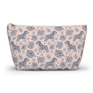 Könnte beinhalten: Eine rosa Reißverschlusstasche mit einem sich wiederholenden Muster aus grauen Schnauzer-Hunden und blauen floralen Elementen. Die Tasche hat einen goldfarbenen Reißverschluss und ein cremefarbenes Innenfutter.