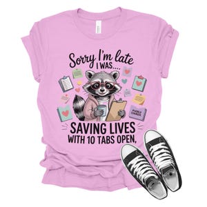 May include: A light pink t-shirt with a graphic of a raccoon wearing glasses and holding a clipboard and coffee cup. The text reads "Sorry I'm late I was... SAVING LIVES WITH 10 TABS OPEN." Black and white sneakers are at the bottom.