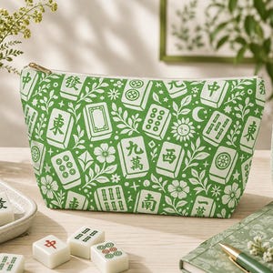May include: A zippered pouch featuring a vibrant green background with a white repeating pattern of mahjong tiles, flowers, leaves, suns, and moons. The rectangular bag has a gold-toned zipper closure along the top edge and a structured shape.